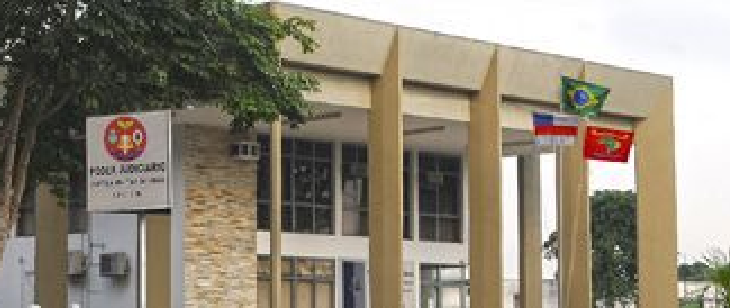 Fachada da 12ª CJM, com jurisdição sobre AM, AC, RO e RR. Edifício térreo com pilares altos e fachada em tons claros. Três bandeiras estão hasteadas em frente ao prédio.