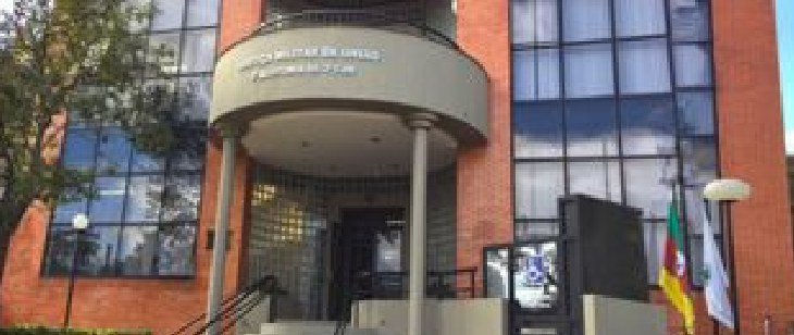 Fachada da 3ª CJM – unidade de Santa Maria. Entrada de um edifício moderno da Justiça Militar da União, com fachada de tijolos aparentes e amplas janelas espelhadas. Duas colunas sustentam uma marquise semicircular com nome institucional. Há bandeiras hasteadas à direita da entrada.