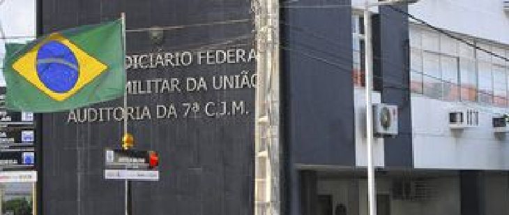 Fachada da 7ª CJM, com jurisdição em PE, AL, PB e RN. Edifício escuro com fachada simples e letreiro institucional visível. Uma bandeira do Brasil está posicionada à frente.