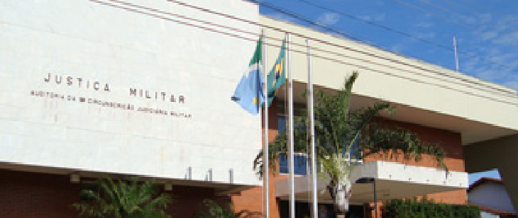 Fachada da 9ª CJM, responsável por MS e MT. Prédio moderno com fachada branca e telhado plano, ladeado por bandeiras e palmeiras. Um abrigo de ponto de ônibus está em frente ao local.